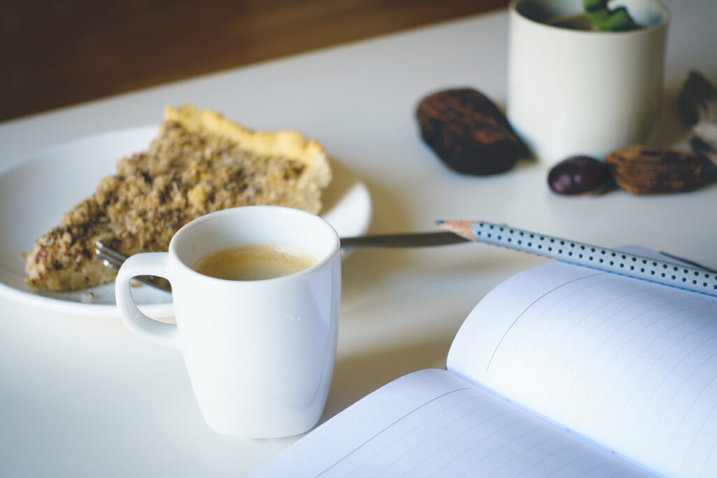 closeup of table with contents on top including pastry, coffee mug, notebook, and pencil